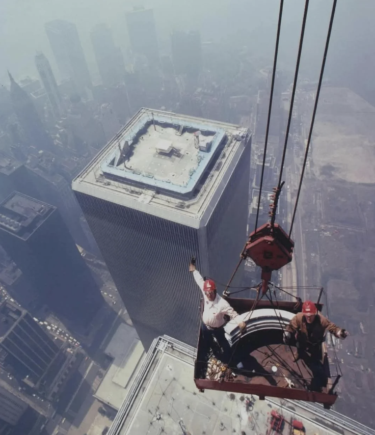 Two construction workers in helmets standing on a suspended platform high above a city, with one waving. A tall skyscraper is visible below them, and hazy buildings and cityscape stretch into the distance under a clear sky.