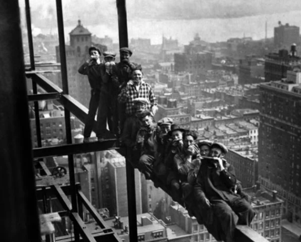 Workers sitting on a steel beam high above a city skyline, eating lunch and smoking. Buildings and cloudy sky are visible in the background, showcasing a construction scene with no safety harnesses.