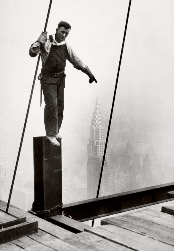 A construction worker in overalls balances on a steel beam high above a city skyline. The Chrysler Building is visible in the foggy background. The worker is holding onto a cable with one hand and pointing down with the other.