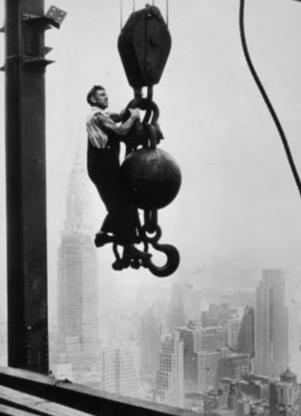 A construction worker in overalls rides on a large hook attached to a crane's cable, high above a city skyline with skyscrapers, including the Chrysler Building, in the background. The scene is black and white, conveying a historical setting.