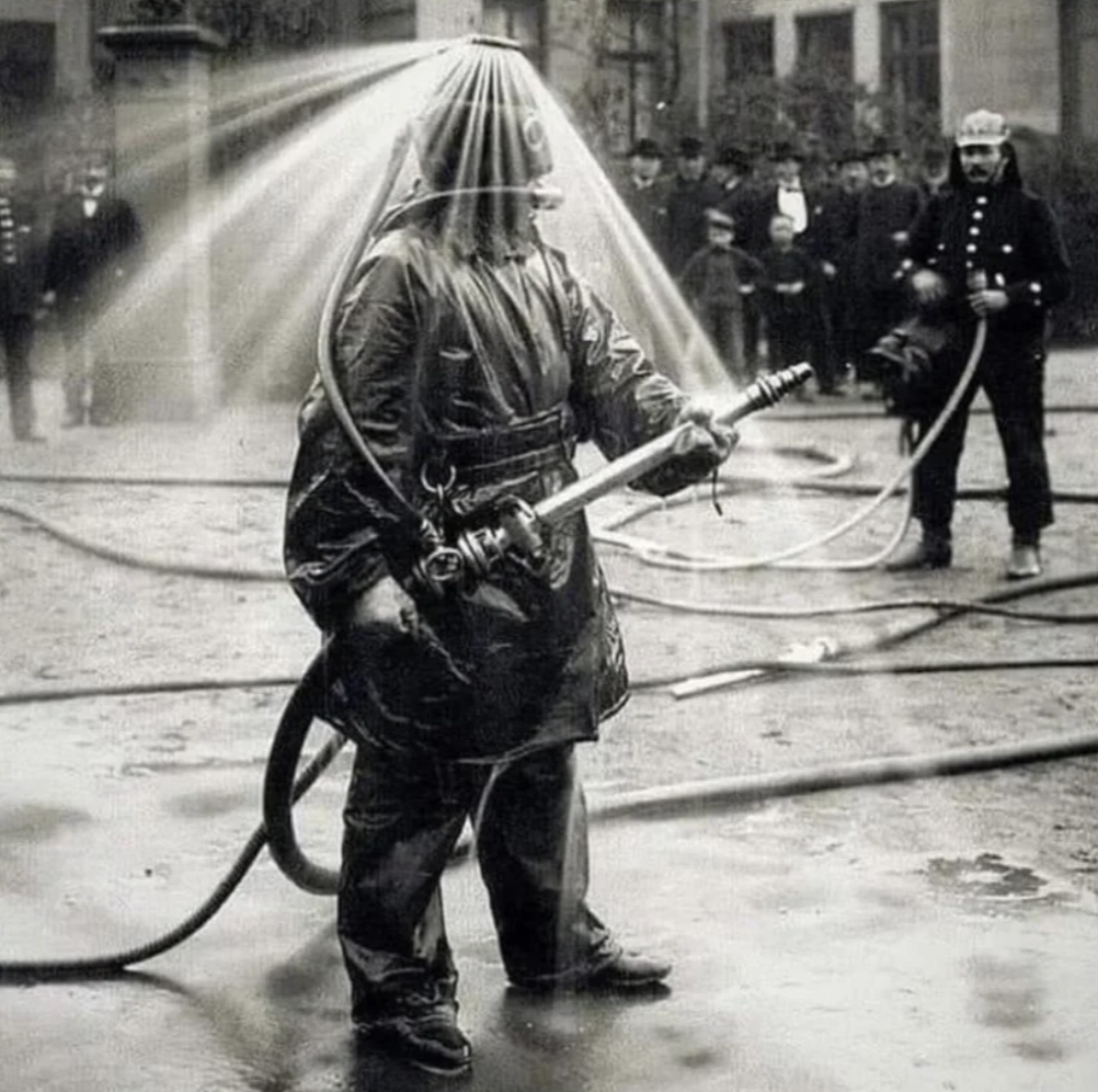 A firefighter in an early 20th-century protective suit sprays water upward, creating a cascading water effect around their head. Onlookers and hoses are visible in the background on a wet street.