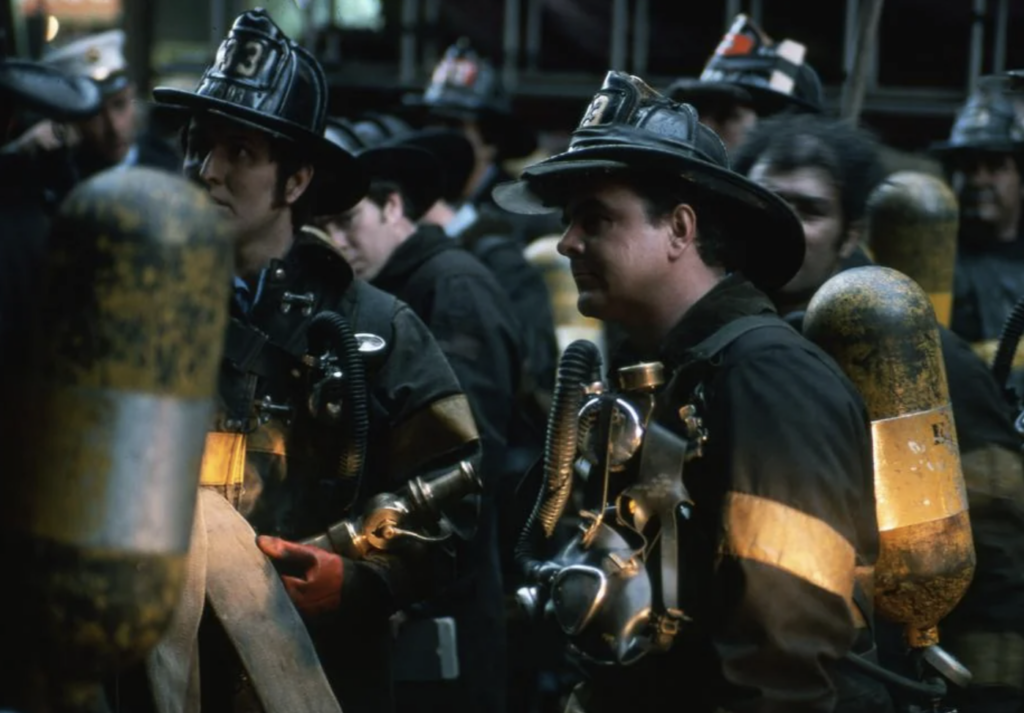 Firefighters in helmets and gear with breathing apparatus stand together, focusing intently. They are surrounded by breathing tanks and appear ready for action in a dimly lit environment, suggesting they are on duty or in a drill.