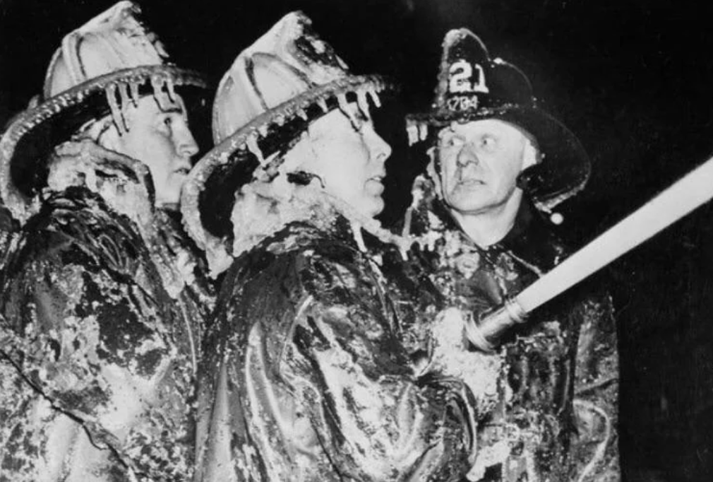 Three firefighters wearing ice-covered gear and helmets spray water from a hose. They work together in challenging freezing conditions, with their coats and helmets glistening with ice as they focus on their task.