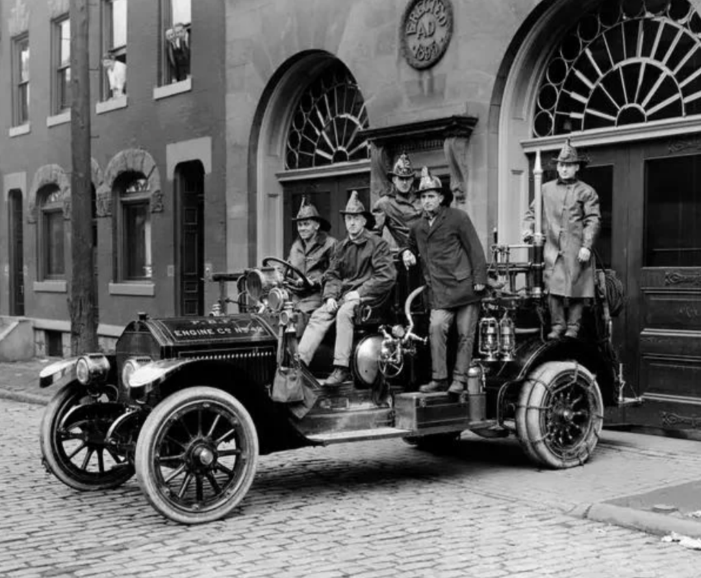 A vintage black and white photo of a group of firefighters posing on an early 20th-century fire engine. The vehicle is parked on a cobblestone street, with a building featuring arched windows in the background.