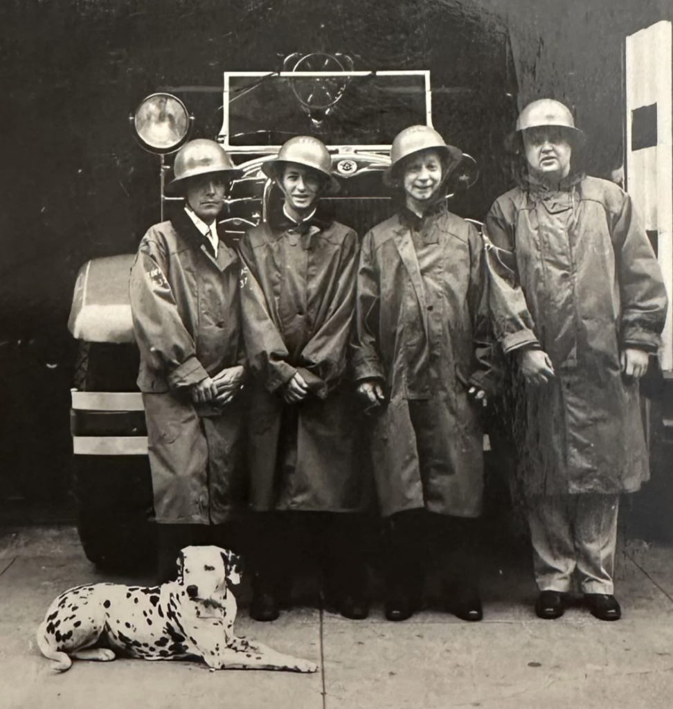 Four people in firefighting gear stand in front of a vintage fire truck. They are wearing helmets and long coats. A Dalmatian lies on the ground in front of them. The photo is black and white, conveying a vintage feel.