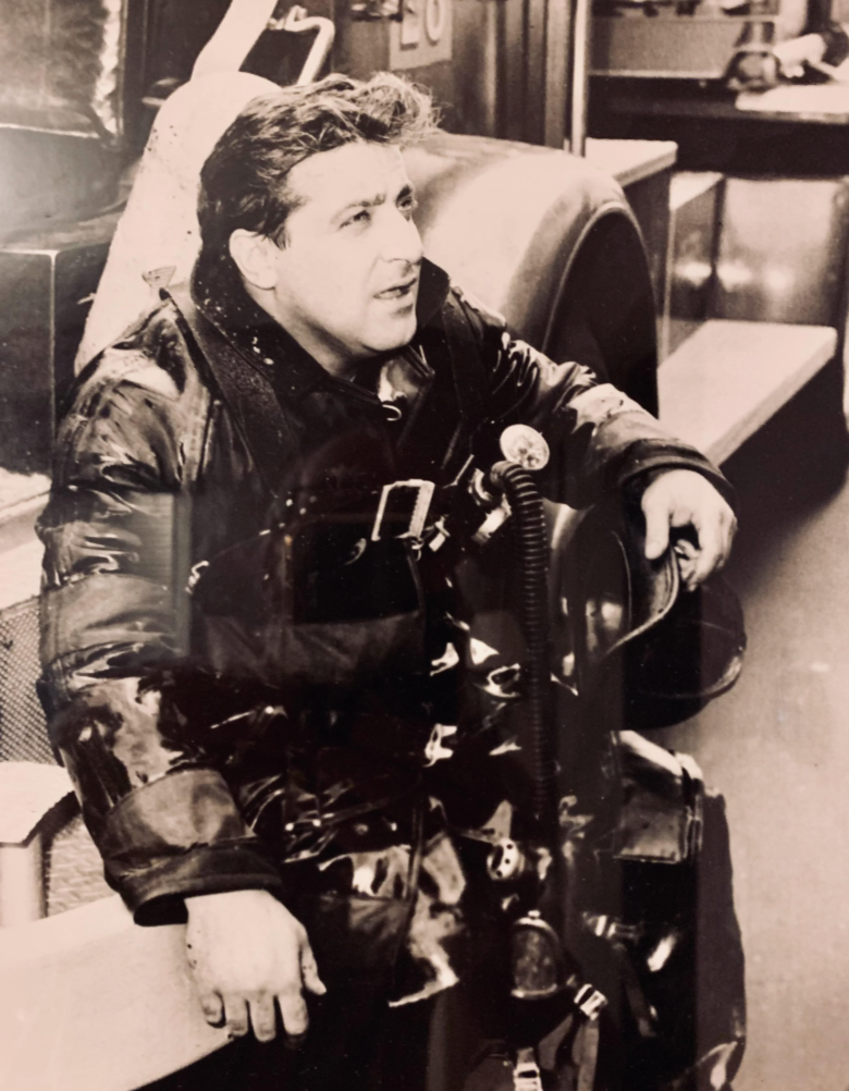 Black and white photo of a firefighter in full gear sitting on the step of a fire truck. The firefighter is looking to the side, wearing a heavy jacket and an air tank. The scene suggests a moment of rest during or after an operation.