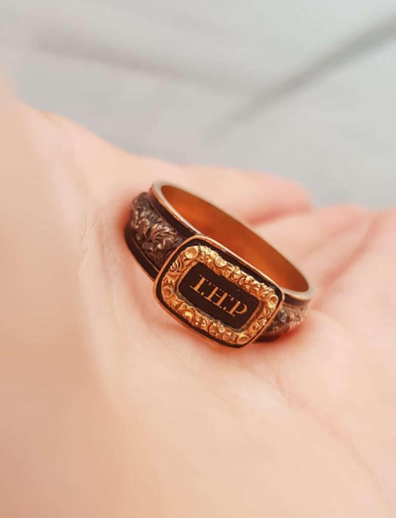 A close-up of a hand holding a vintage gold ring with an ornate design. The ring features an engraved rectangular plate with the initials "LHP." The surrounding band has intricate floral detailing.