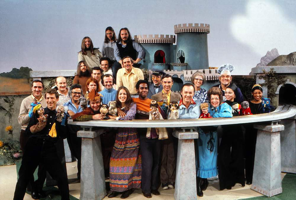 A diverse group of people, some holding puppets, pose in front of a whimsical castle set. They are dressed in colorful 1970s attire, with a mix of men and women smiling at the camera. The background features a castle and painted scenery.