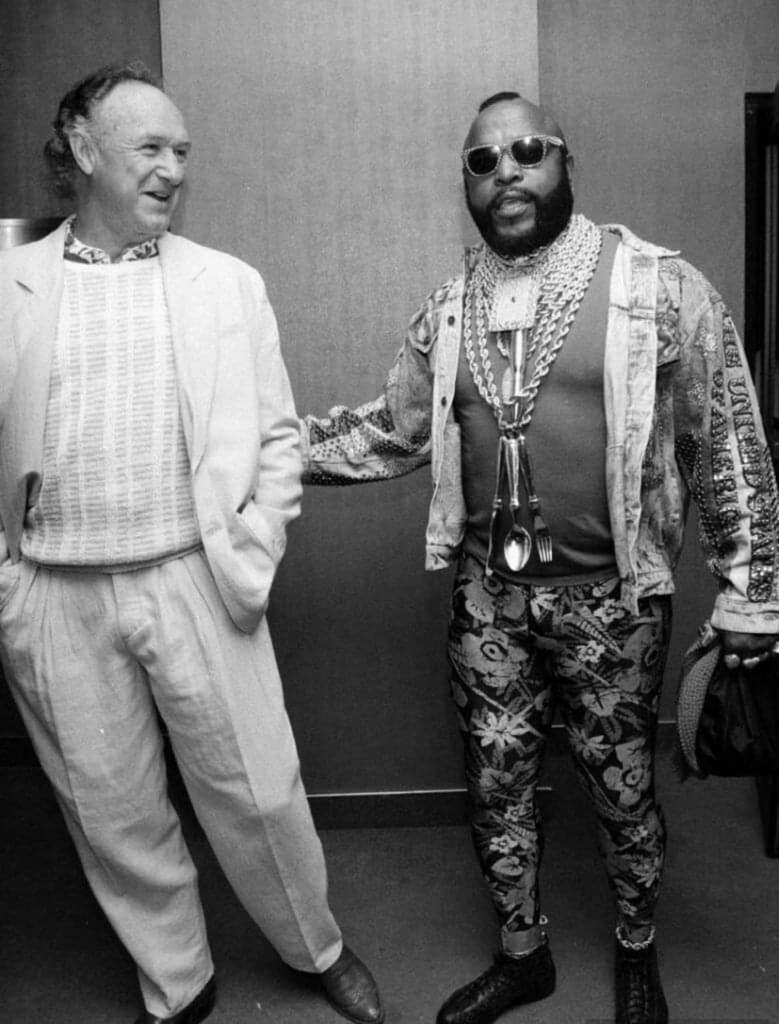 A man in a light suit and patterned sweater smiles while standing next to another man wearing sunglasses, a patterned jacket, leggings, and multiple large necklaces, including spoons, in a stylish and bold outfit.