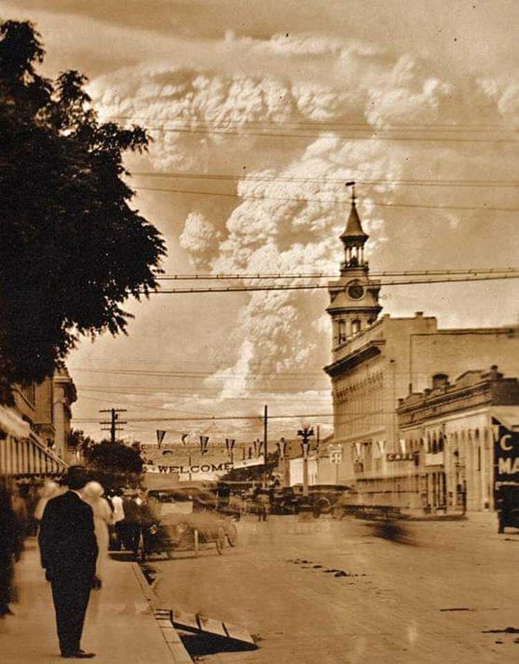 Sepia-toned image of a historic street scene with vintage cars. A clock tower rises in the center, and banners hang overhead. Large clouds billow in the sky, and blurred pedestrians walk on the sidewalks. A man stands in the foreground.