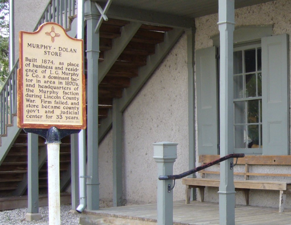 A historical marker stands in front of a gray building with wooden steps and a porch. The sign details the Murphy-Dolan Store, built in 1874, highlighting its role in local business, county government, and as a judicial center for 33 years.