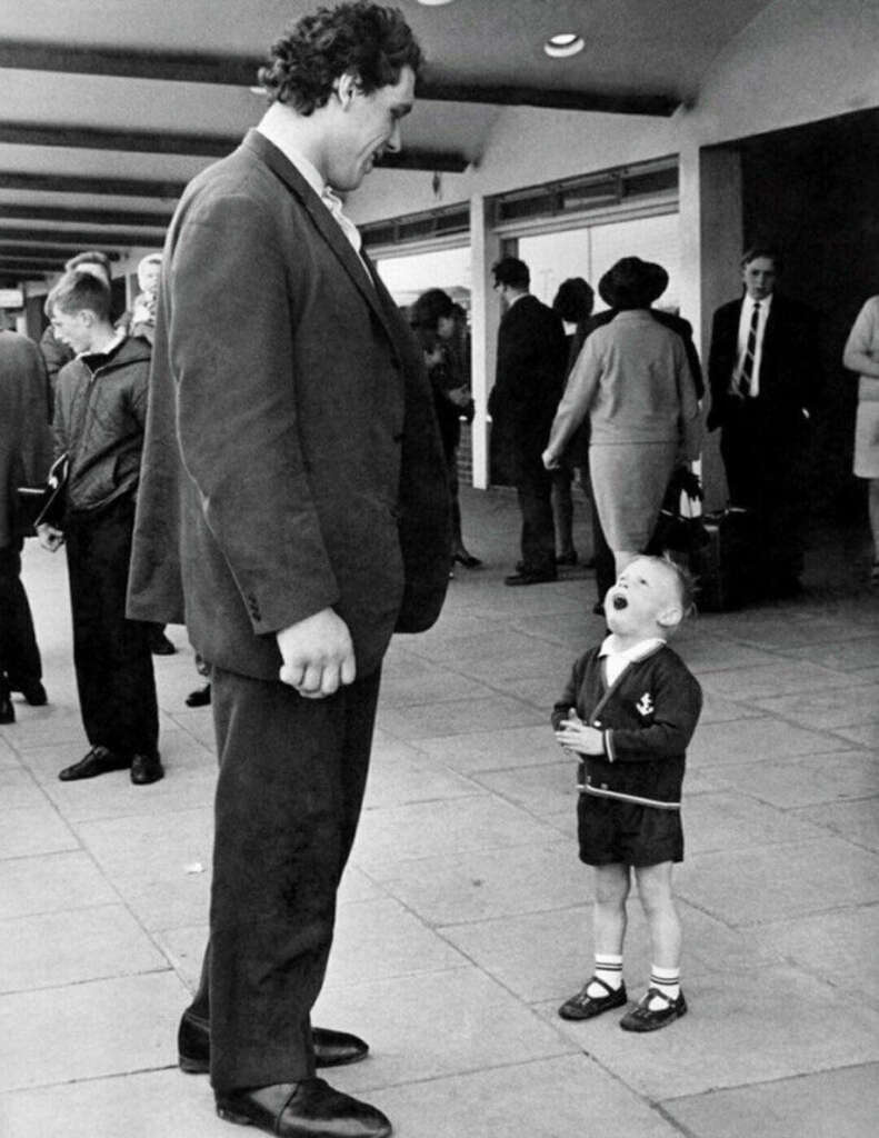 A tall man smiles down at a small child who is laughing and looking up at him. They are outdoors, surrounded by people walking by. The child is wearing a dark jacket and shorts, while the man is in a suit.