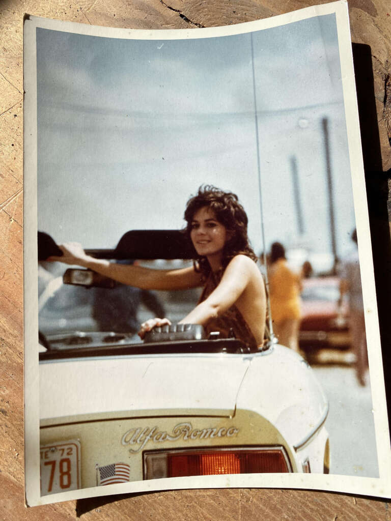 A person with long hair smiles while driving a vintage Alfa Romeo convertible. The car has a U.S. flag sticker and a partially visible license plate. The background shows a blurred outdoor setting with other people and vehicles.