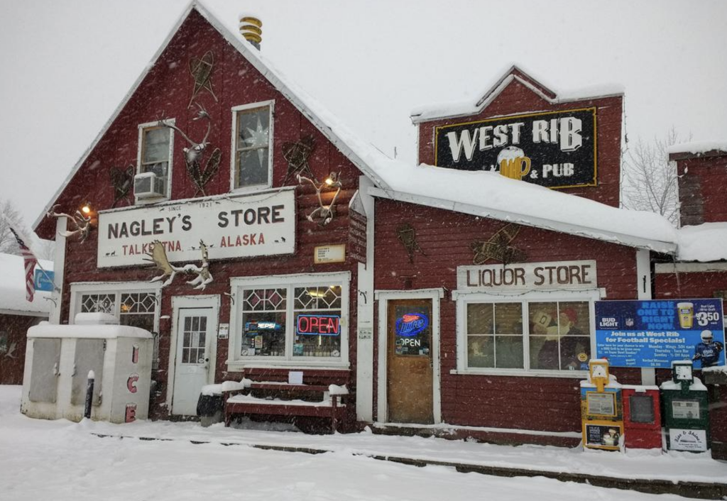 A rustic red building covered in snow, featuring "Nagley's Store" and "West Rib Pub" signs. It also serves as a liquor store. Several antlers decorate the exterior. There are snow-covered benches and a sign advertising food specials outside.