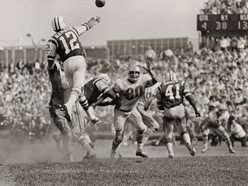 A vintage black-and-white photo of an American football game. A quarterback wearing number 12 leaps to throw the ball as defenders approach. Other players in numbered jerseys pursue. A crowded stadium filled with spectators is in the background.