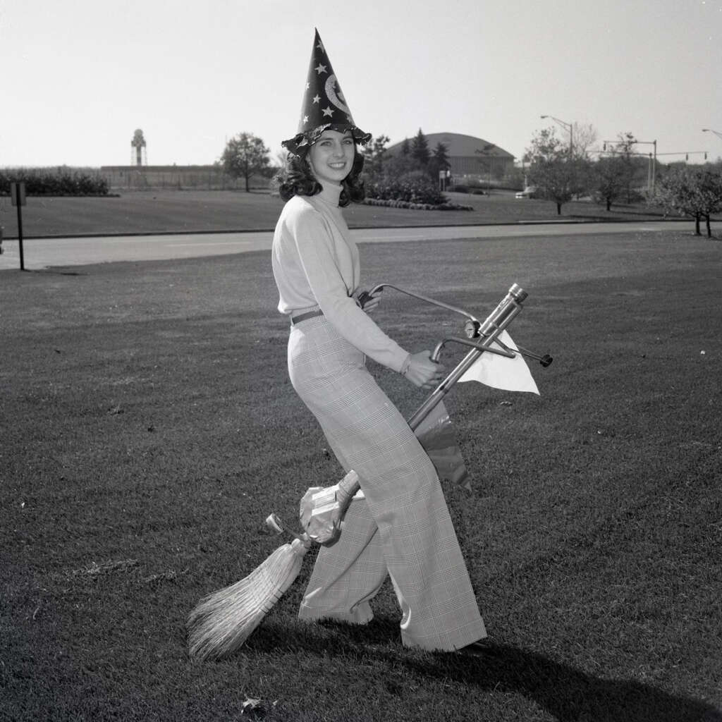 A person wearing a star-patterned wizard hat and light-colored clothing sits on a decorative broomstick shaped like a bicycle, positioned on a grassy field with trees and a building in the background. The image is in black and white.