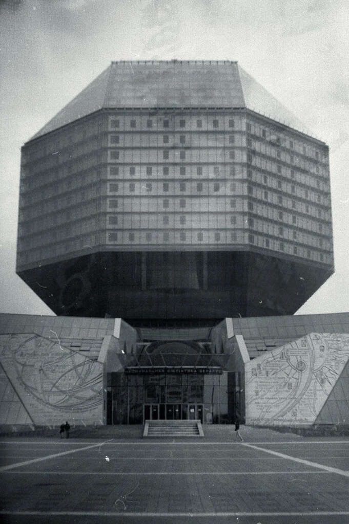 A black and white photo of a building with a large, geometric, diamond-shaped structure elevated above a wider base. The facade features numerous windows, and the entrance is at the center of the lower section. The sky is overcast.