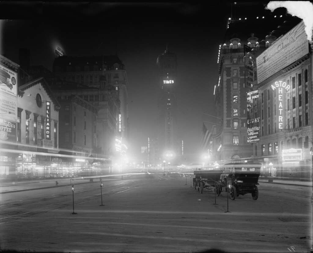 Black and white nighttime photo of Times Square, New York, in the early 1900s. The scene is illuminated by bright signs and lights. An antique car and a carriage are visible on the street, with buildings on either side.