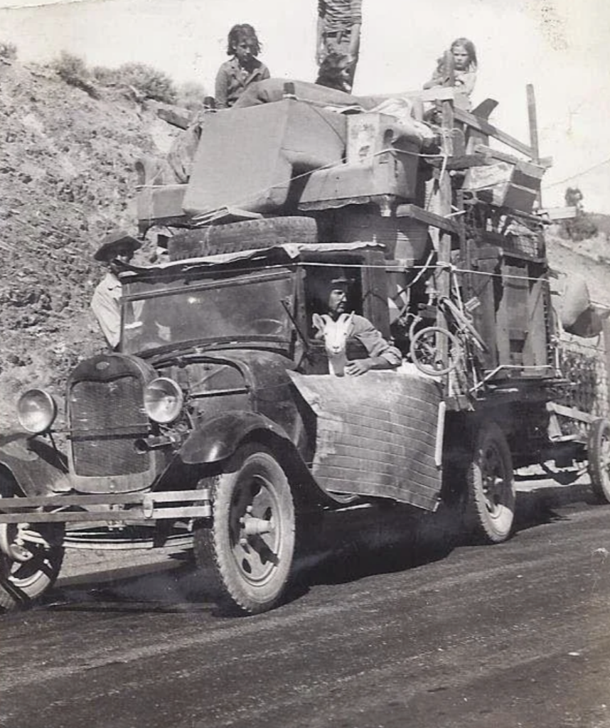 A black and white photo of an old truck loaded with furniture and household items. It has makeshift wooden structures. Two people sit in the front seat and children are visible among the items. The truck is on a paved road with a hilly background.