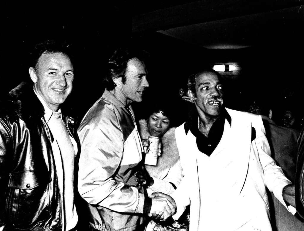 A black and white photo of four people smiling and interacting at an event. The person on the left wears a leather jacket, and the person on the right wears a white suit. The middle group includes one person holding a drink.