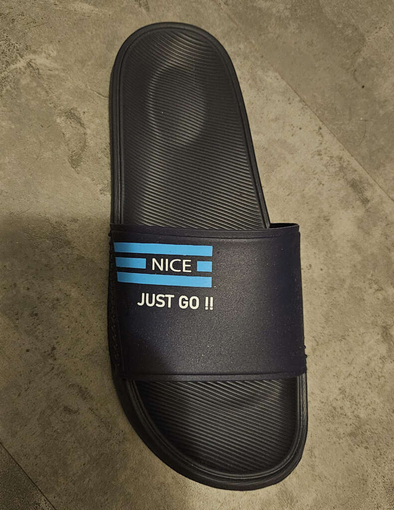 A single black slide sandal with blue stripes and the words "NICE JUST GO!!" on the strap. The sandal is on a textured gray surface.