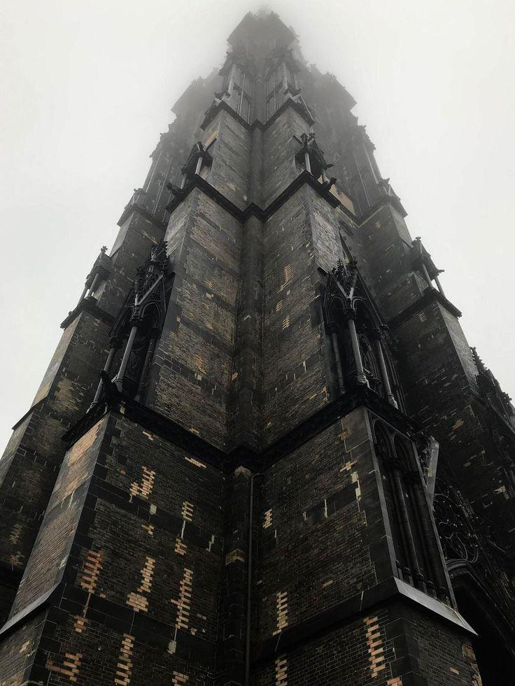 A towering Gothic-style building with an intricate facade rises into a dense layer of fog. The stone structure features dark bricks and ornate detailing, emphasizing its height and grandeur against the misty backdrop.