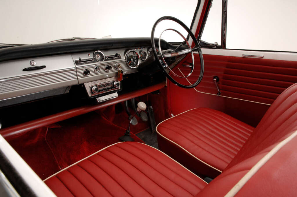 Interior of a vintage car with a red and white color scheme. Features include red leather seats, a classic steering wheel, and an old-fashioned dashboard with analog dials and a radio. The gear shift is mounted on the floor.