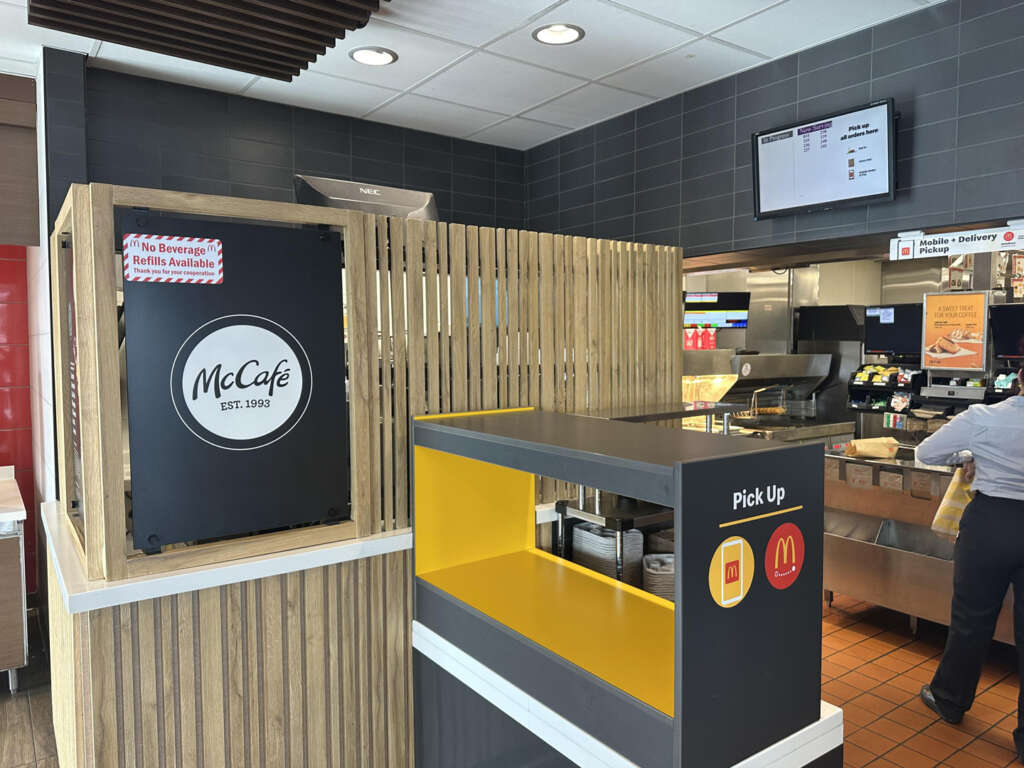 A McDonald's interior showing a McCafé area with a sign indicating "No Beverage Refills Available." The counter has a pickup area with a yellow and gray sign. An employee is in the background, and a digital display is mounted on the wall.