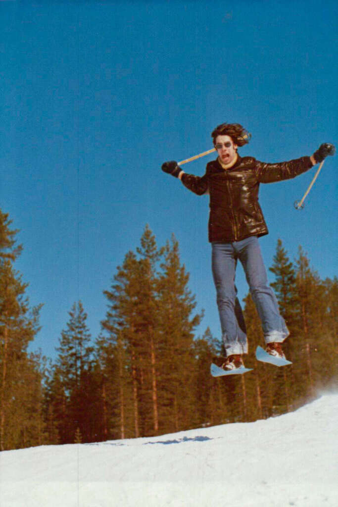 A person in a black jacket and jeans is mid-air while skiing, holding ski poles. They are jumping off a snowy slope with tall pine trees in the background, under a clear blue sky.