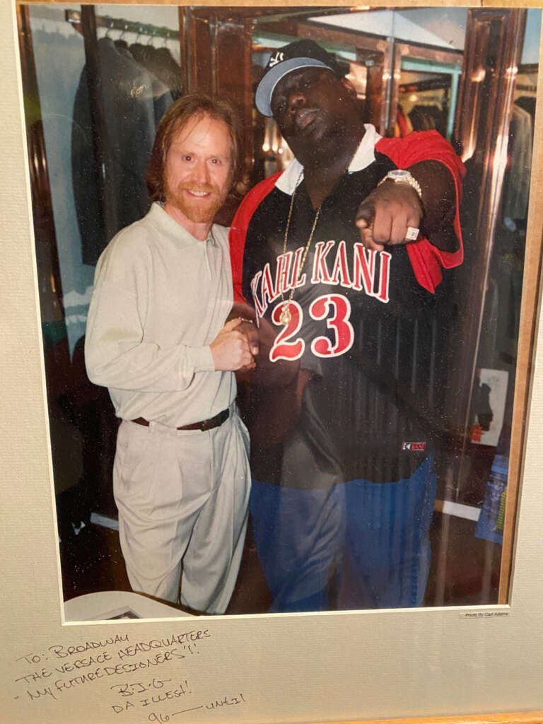 Two men posing together for a photo. The man on the left has long hair and a beard, wearing a light-colored outfit. The man on the right is wearing a cap and a jersey with "Karl Kani 23," pointing towards the camera. Handwritten text is at the bottom.