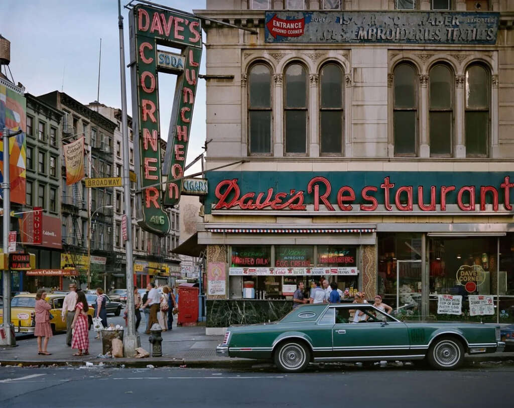 Street scene featuring Dave's Corner Luncheon and Dave's Restaurant on a city block with vintage signage. A green classic car is parked in front. People are walking on the sidewalk, with various shops and signs visible in the background.