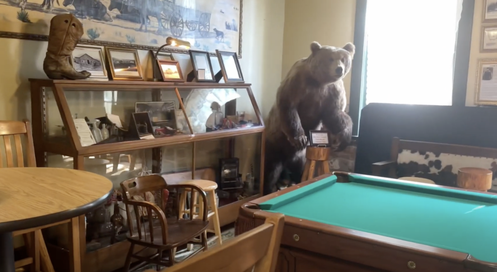 A room with a pool table, wooden chairs, and a display cabinet filled with framed photos and artifacts. A large taxidermy bear stands beside the cabinet. A large painting of old Western imagery hangs above the display.