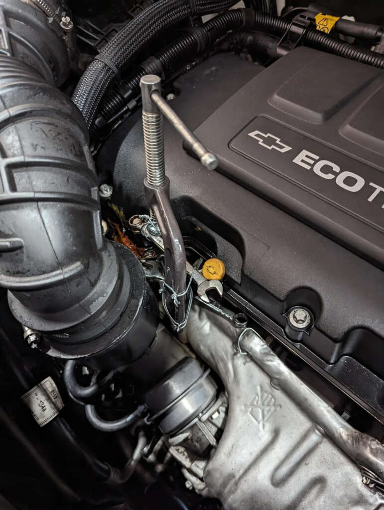 Close-up of a car engine showcasing a dipstick inserted next to the oil cap. The engine cover features the Chevrolet logo and "ECOTEC" branding. Various pipes and components are visible around the engine.