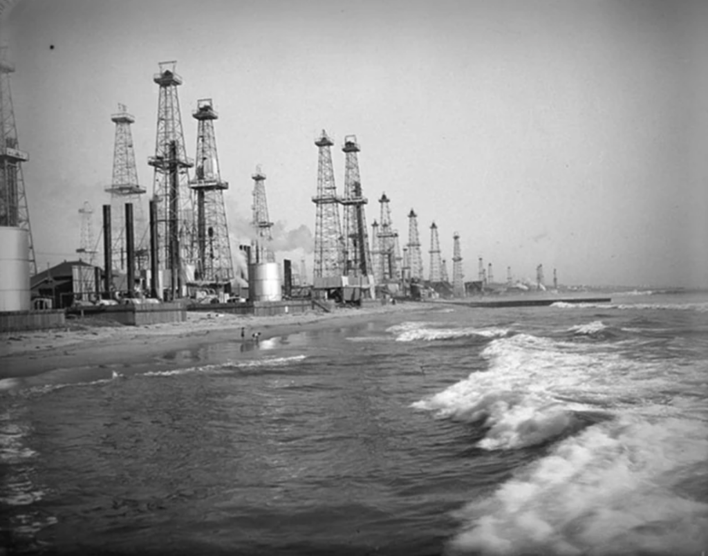 A black-and-white photograph showing a row of numerous oil derricks along the shoreline. Waves are visible in the foreground, and the sky is clear. The derricks are evenly spaced and extend into the distance.