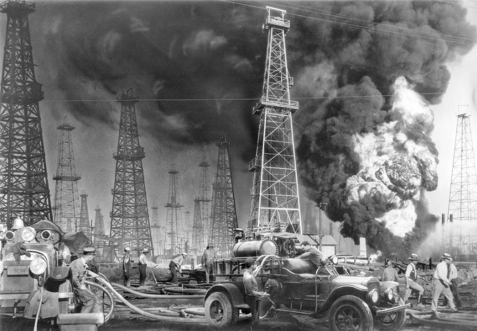 Black and white photo of firefighters battling a large oil fire at an oil field. Smoke billows behind multiple oil derricks. Fire trucks and equipment are in the foreground, with several firefighters in action.