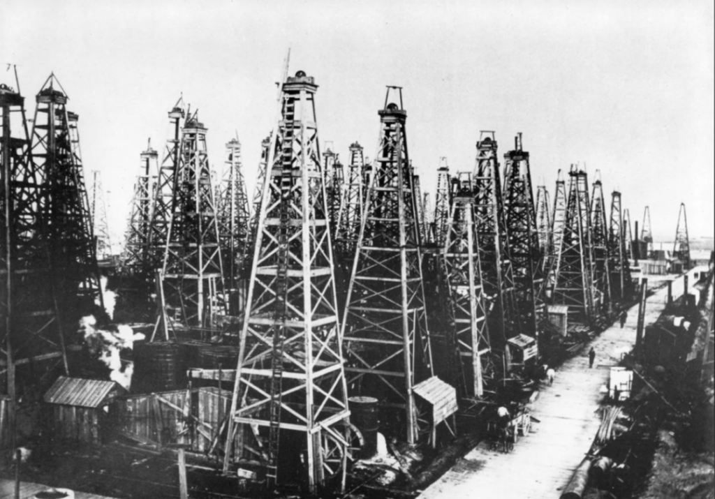 Black and white photo of numerous wooden oil derricks closely packed together, with small sheds and buildings at their bases. A dirt path runs alongside them, and the sky is clear. The scene suggests an early 20th-century oil field.