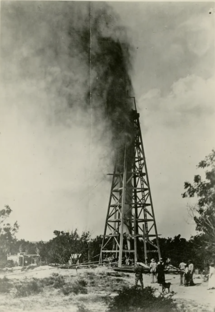 An old black-and-white photo of an oil derrick with oil gushing out. Several people stand at the base, observing the scene. Trees and small structures are in the background under a cloudy sky.