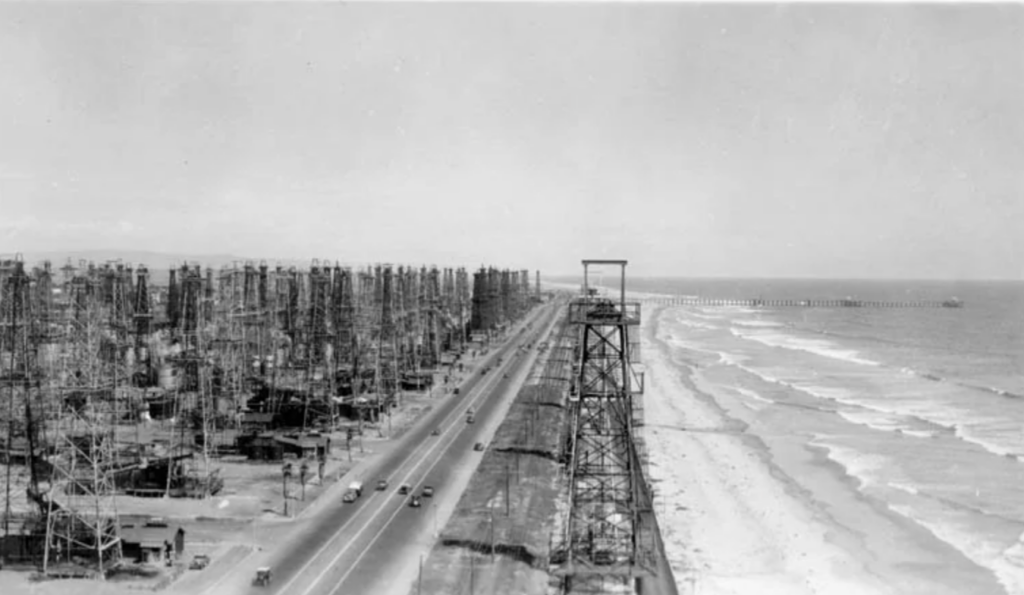 Vintage black and white photo of an old highway running parallel to an ocean shoreline. Numerous oil derricks line the road on the left side, stretching into the distance, with the sea visible on the right side.