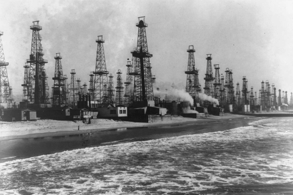 Black and white image showing numerous oil derricks lined up along a beach, with waves in the foreground. Smoke rises from structures among the derricks, indicating industrial activity in a coastal setting.