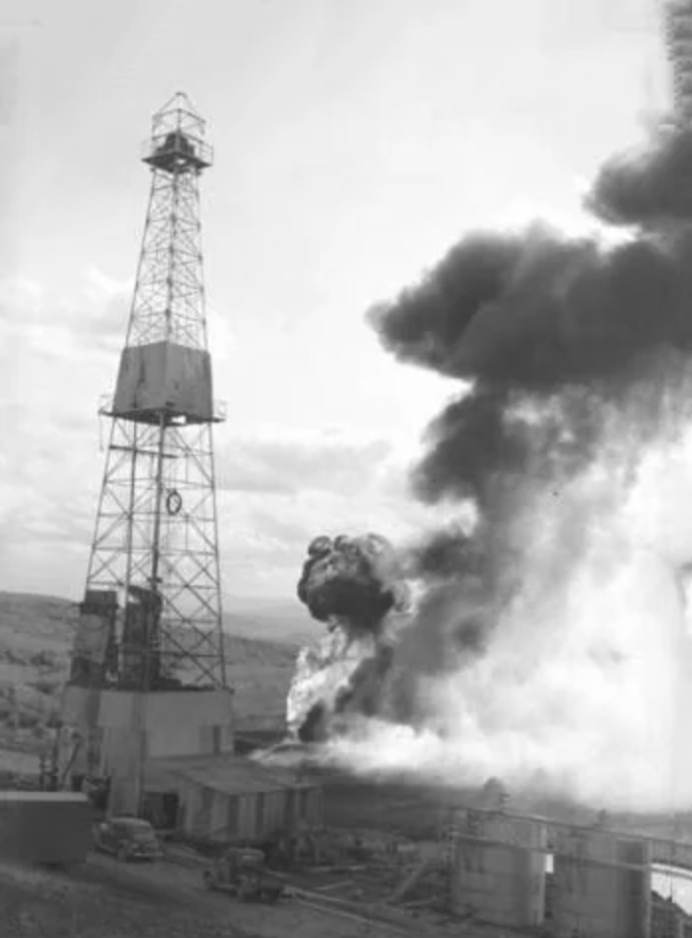 Black and white image of a tall oil derrick with thick, dark smoke billowing nearby. The landscape is flat, with open fields in the background. Structures and vehicles are visible at the base.