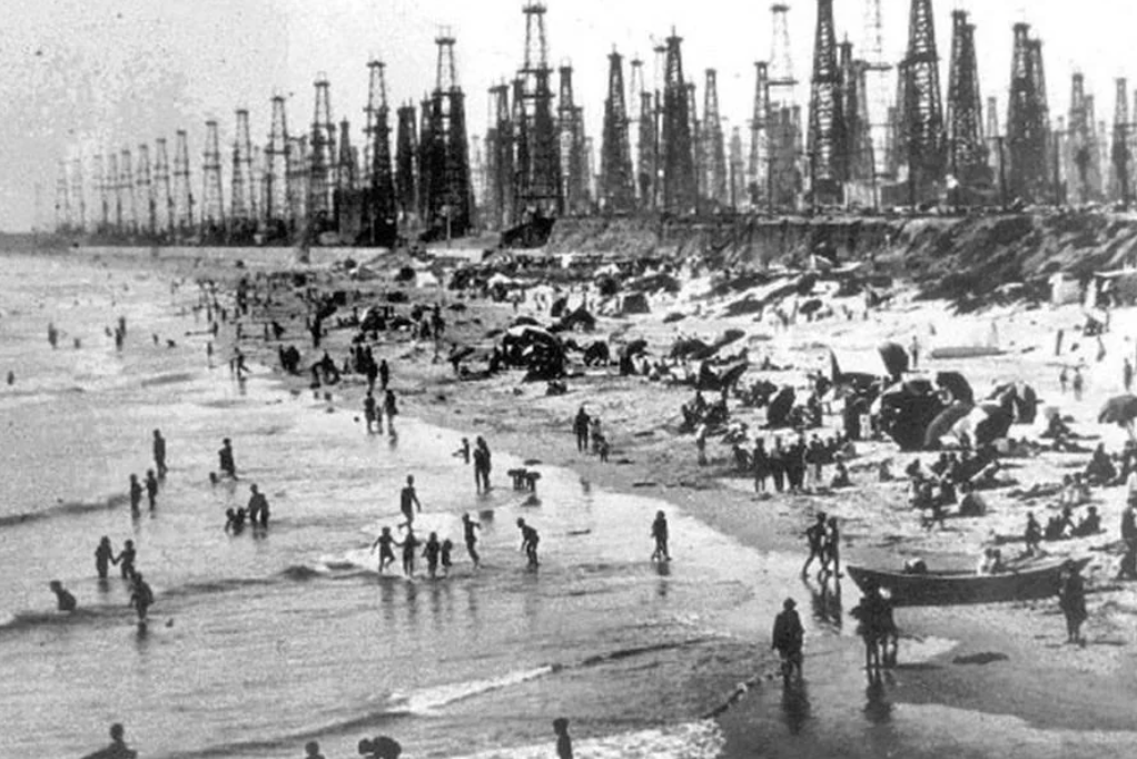 Black and white photo of a crowded beach lined with multiple tall oil derricks. People are swimming, walking, and gathering along the shoreline. The scene has an industrial backdrop contrasted with recreational beachgoers.