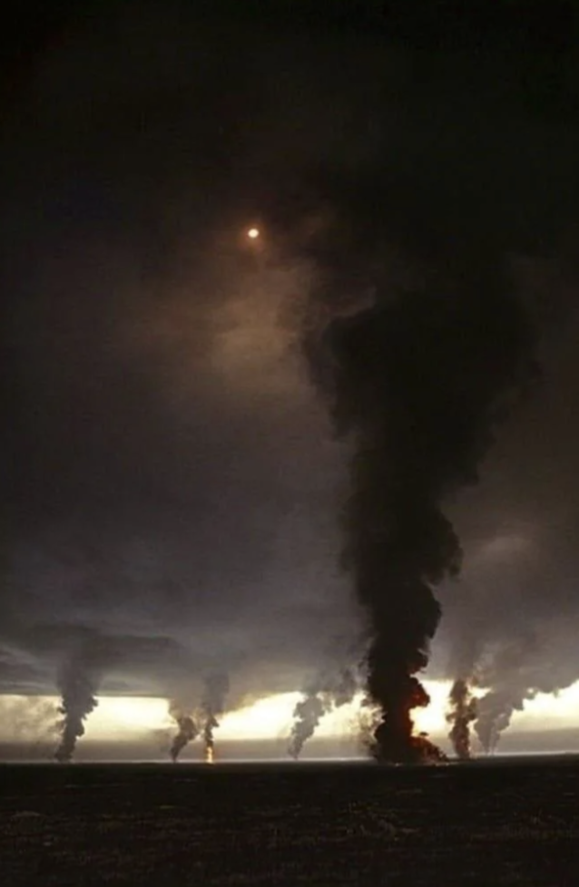 Thick black smoke billows into the sky in front of a dimly lit horizon, partially obscuring the sun. The scene appears dramatic and intense, with multiple plumes of smoke rising from ground level.