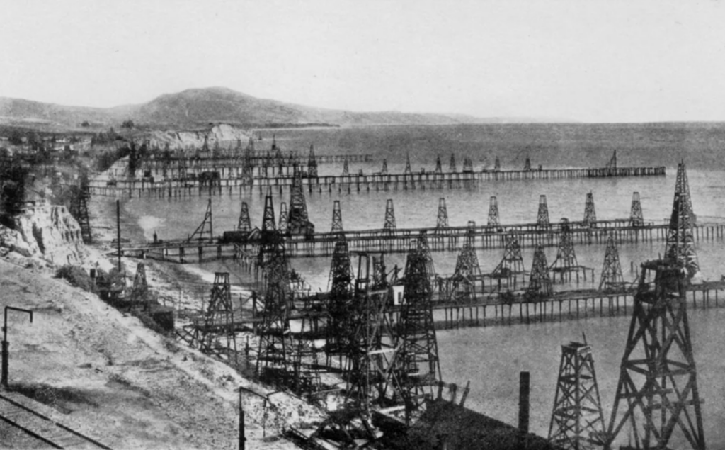 A historical black-and-white photo showing numerous oil derricks on raised platforms extending over a body of water along a coastline. Hills are visible in the background, under a clear sky.