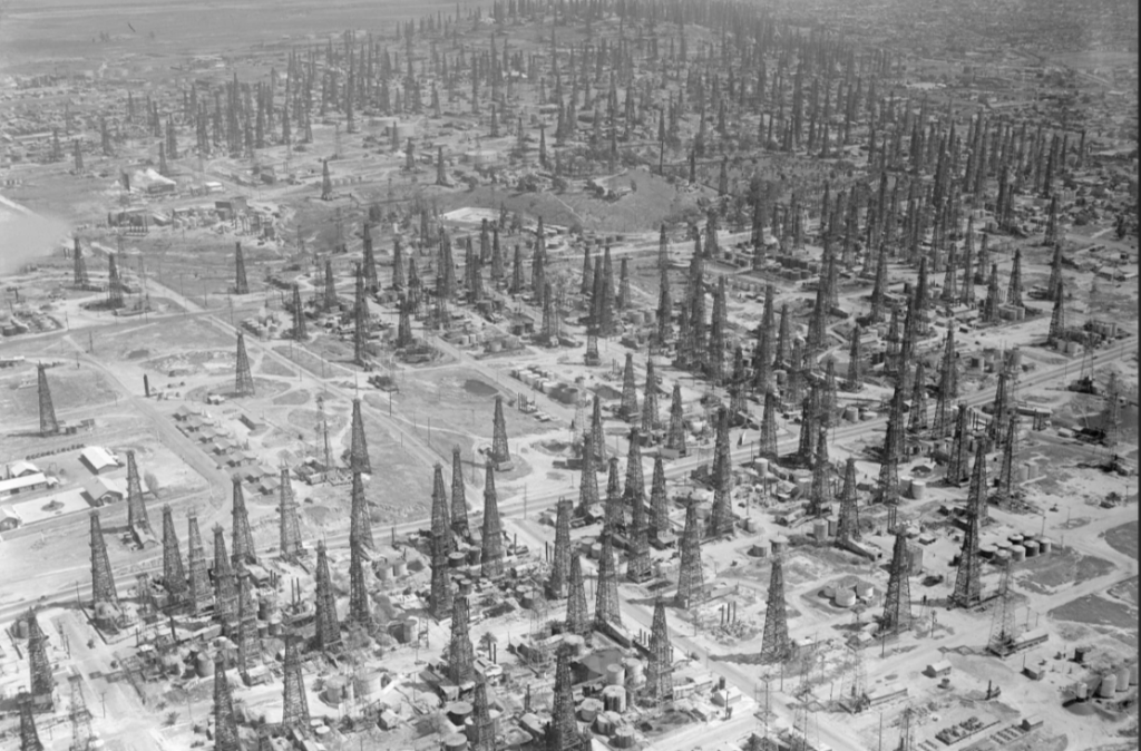 Black and white aerial view of a densely packed oil field, featuring numerous oil derricks spread across a vast landscape. The image captures an industrial scene with a network of roads connecting the structures.