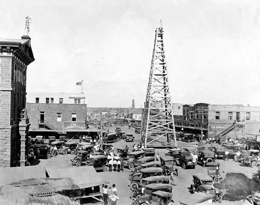 Black and white photo of a bustling 1920s street with numerous vintage cars and people. A large wooden oil derrick stands prominently in the center. Surrounding buildings include shops and a hotel, with signs visible. The scene is lively and crowded.