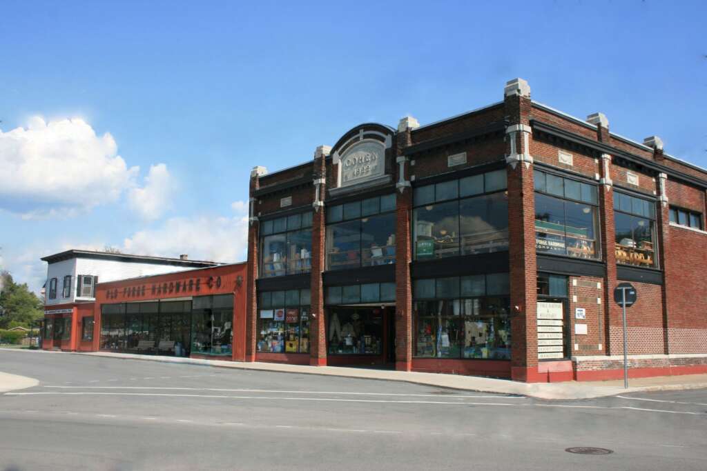 A two-story brick building with large glass windows and a sign reading "York Emporium" stands at a street corner under a blue sky. Adjacent is a smaller white building with decorative elements. Sidewalks run alongside both structures.