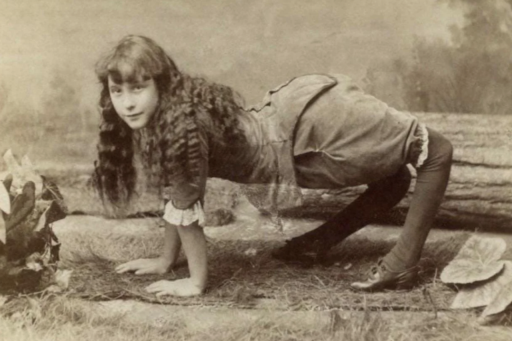 A sepia-toned vintage photograph of a young girl with long curly hair, dressed in Victorian-era clothing and boots. She is posed playfully on her hands and feet, with her knees bent, against a rustic backdrop of logs and foliage.