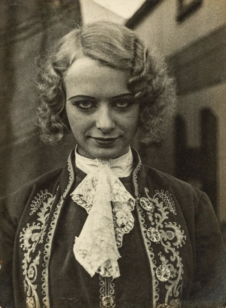 A black and white portrait of a person with curly hair wearing an ornate, embroidered jacket and a lace cravat. The background features blurred buildings, giving a vintage feel to the image.