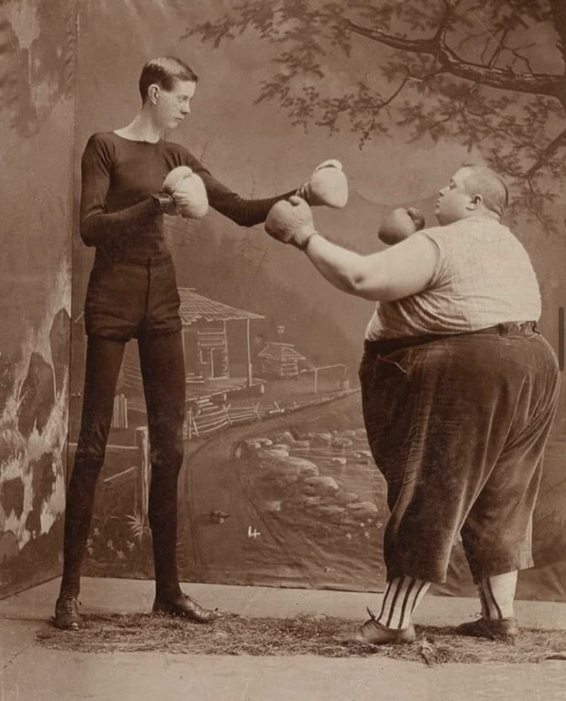 A vintage sepia photo of two men boxing. One is tall and thin, in a black outfit, while the other is shorter and heavier, wearing dark pants and a striped shirt. They face each other on a rustic studio backdrop.