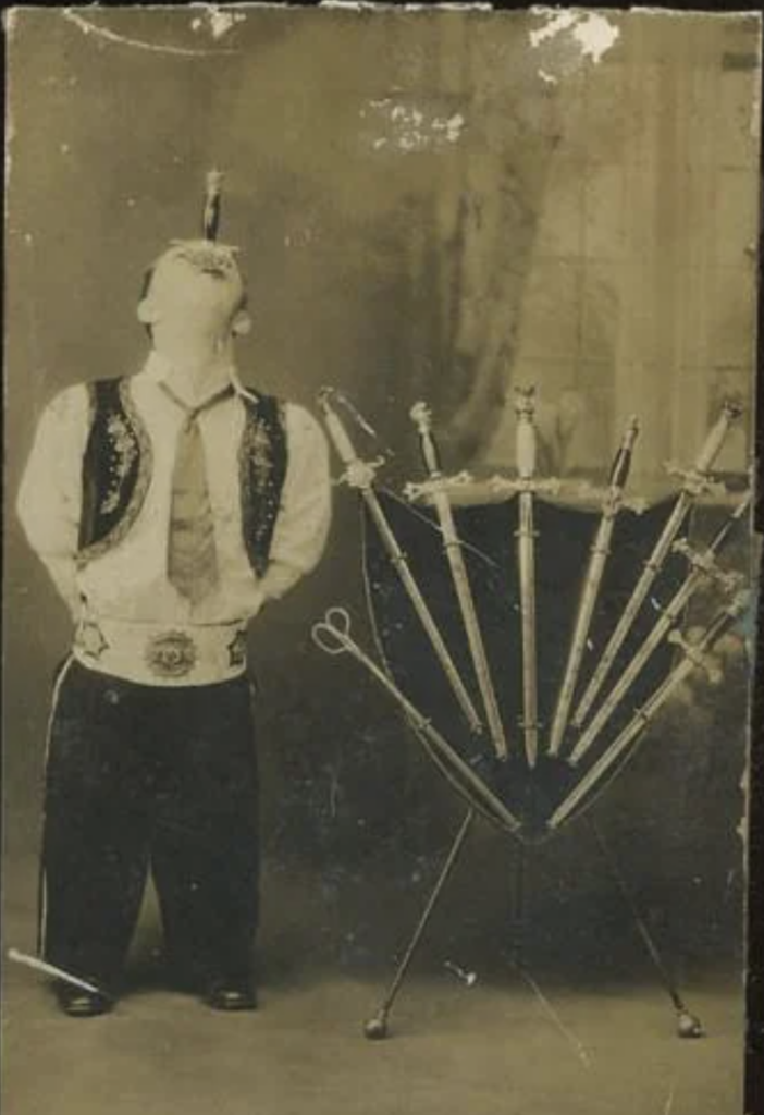 A vintage photograph shows a performer swallowing a sword while standing next to an umbrella stand holding several swords. The performer is dressed in traditional attire with a vest and wide belt. The scene is set against a simple backdrop.